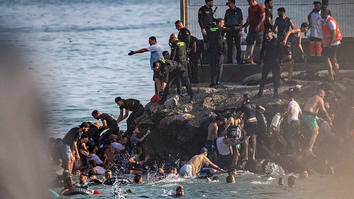 Migrants swimming and climbing onto a rocky breakwater in Ceuta as Spanish Guardia Civil officers watch from above.