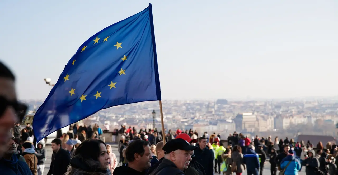 A blue European Union flag with yellow stars held up in front of a crowd, with a city skyline in the background.