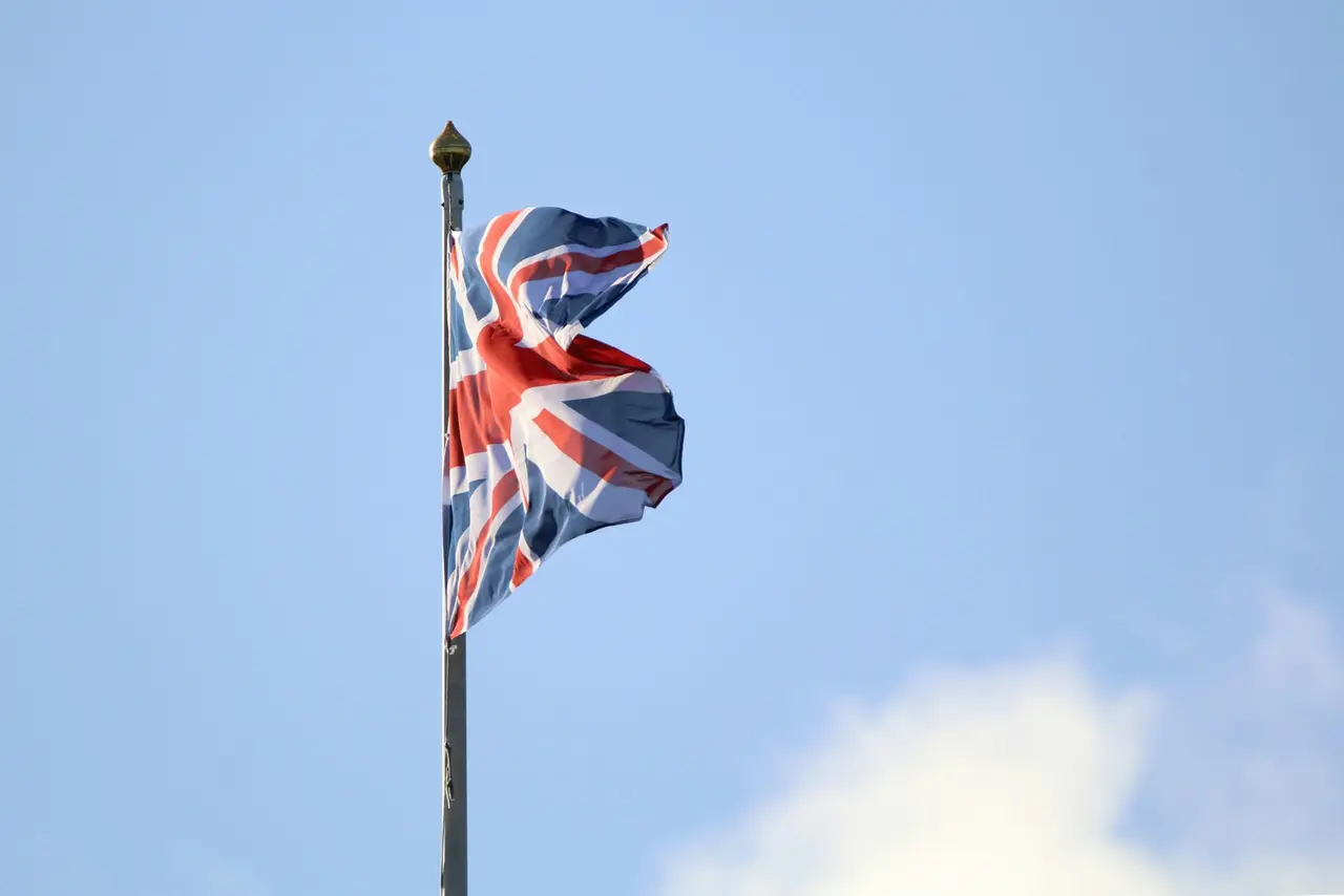 A close-up photograph of the British Union Jack flag waving from a tall flagpole against a bright blue sky with light clouds.